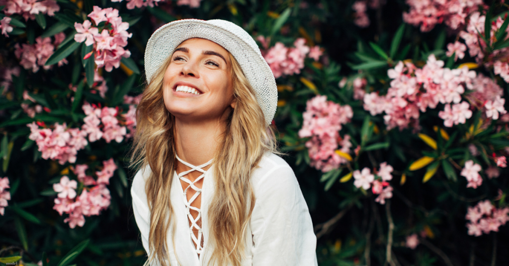 Young woman with long blonde hair and a hat smiling broadly in front of bushes in bloom with pink flowers