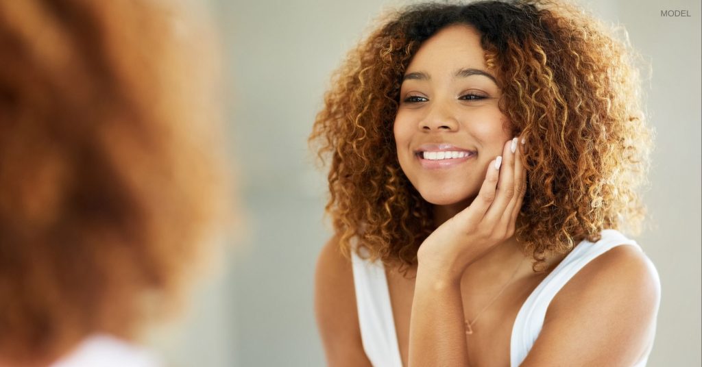 Woman with clear skin (model) looking in the mirror with a hand held to her face.